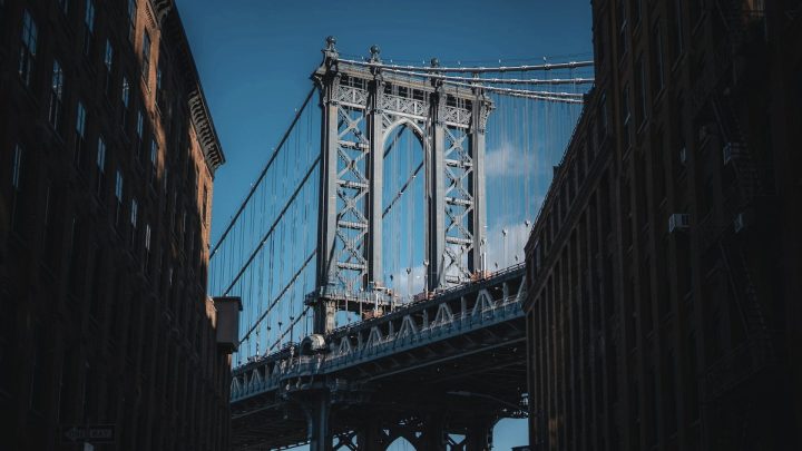 View of Brooklyn Bridge in New York City framed between two tall brick buildings on a clear blue day.