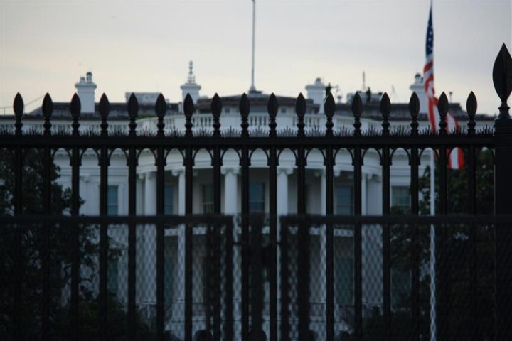 White House seen behind a black iron fence, with a U.S. flag visible in the background.